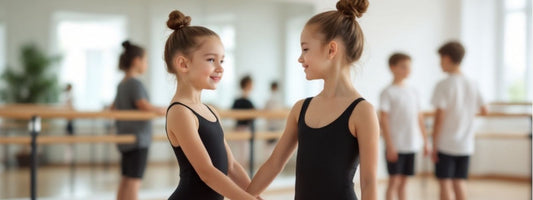 Two young girls in black ballet leotards smiling and holding hands in a dance studio, with other children practicing in the background