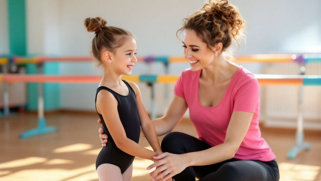 A smiling dance teacher kneeling and encouraging a young girl in a black leotard inside a bright and colorful dance studio with ballet barres in the background